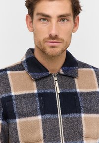 Wool plaid jacket featuring beige, navy, and light blue squares, a zip front, and a collar with a soft texture.