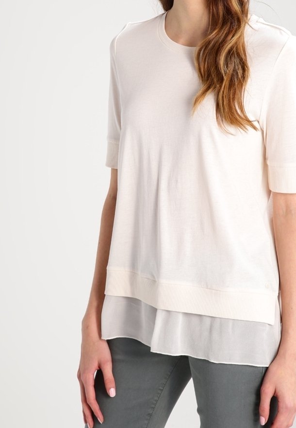 Jasnokremowa koszulka z krótki rękawami i warstwowym dołem z przejrzystego materiału. Posiada okrągły dekolt i luźny krój. Miękka tekstura.