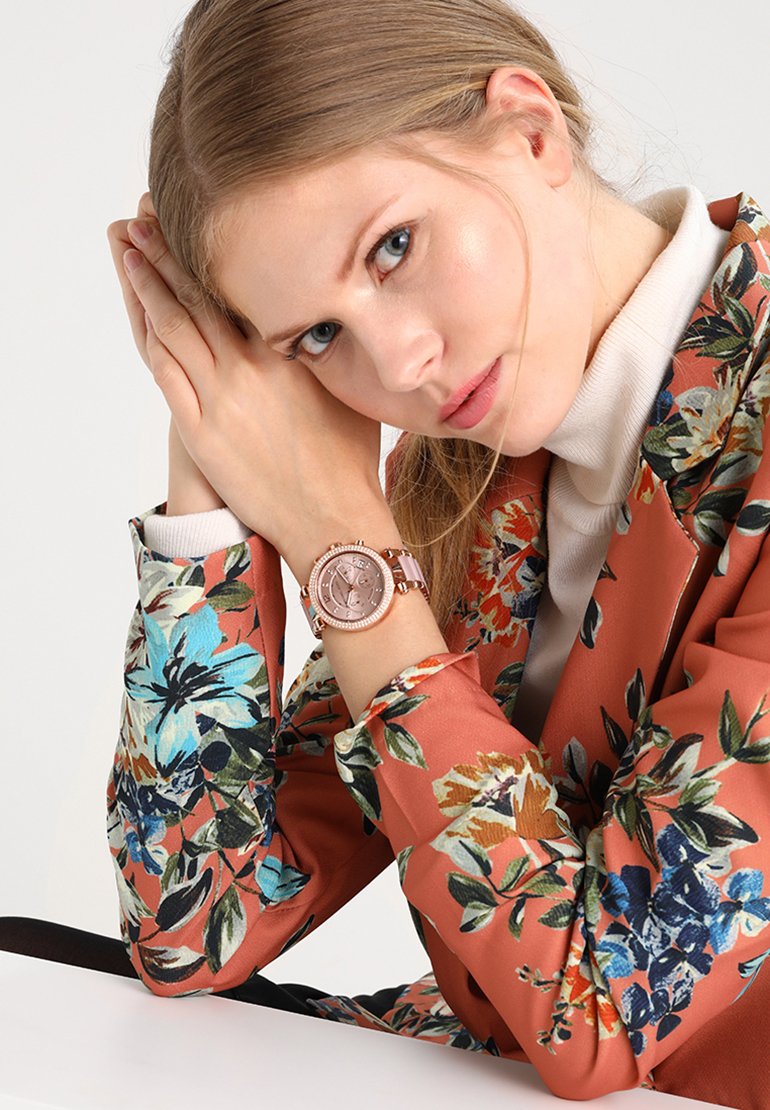 Žena s modrýma očima spočívající hlavou na sepjatých rukou, oblečená v květovaném hnědém saku a na zápěstí má růžově zlaté hodinky.