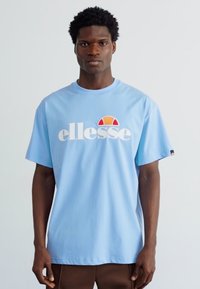 Gaiši zils viegls kokvilnas T-krekls ar lielu centrētu balto "ellesse" logotipu, ap kuru ir sarkans un oranžs pusaplis. Īsās piedurknes.