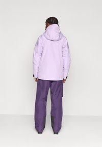 Chaqueta impermeable morada con capucha, combinada con pantalones morados de corte holgado que tienen detalles negros en los tobillos. Textura suave.