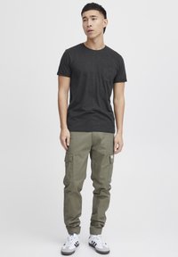 T-shirt noir à manches courtes avec une poche sur la poitrine, associé à un pantalon cargo vert olive doté de poches latérales, et des baskets blanches avec des accents noirs.