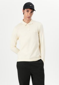 Polo à manches longues crème avec un col, deux boutons et un petit logo en patch. Porté avec un pantalon noir et une casquette noire. Texture lisse.