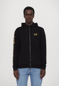 Zwarte zip-up hoodie van een zachte stof, met een geel EA7-logo op de borst en lange mouwen met logo-tekst langs één arm.