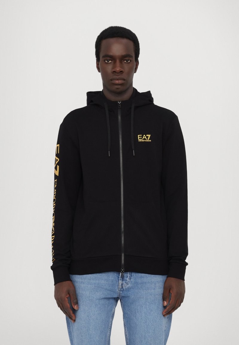 Zwarte zip-up hoodie van een zachte stof, met een geel EA7-logo op de borst en lange mouwen met logo-tekst langs één arm.