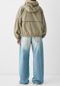 Osoba ubrana w beżową kurtkę z kapturem i luźne, jasnoniebieskie, wyblakłe dżinsy, widoczna od tyłu na jednolitym białym tle.