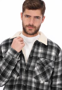 Chaqueta de camisa a cuadros en blanco y negro con cuello de piel sintética blanca, dos bolsillos delanteros, tela texturizada y cierre frontal con botones.