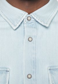 Camicia in denim blu chiaro con colletto, placca centrale con bottoni simili a perle e due tasche sul petto. Il tessuto ha un motivo a trama.