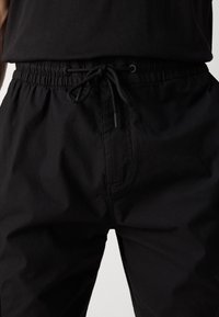 Gros plan d'un pantalon noir avec une ceinture élastique et un cordon noué, porté avec un haut noir.