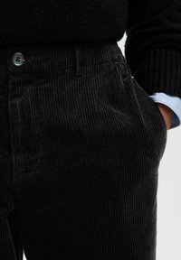 Mustad corduroy püksid, millel on nupp ja küljetaskud, kaasas must kampsun ja sinise triibuga särgi varrukas on nähtav ühel randmel.