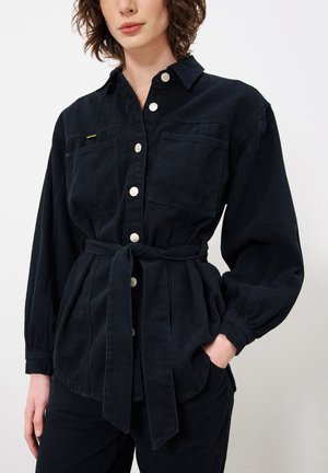 Zwarte denim shirt met knoopsluiting aan de voorkant, twee borstzakken, strik taille en lange mouwen. Heeft een zachte textuur en zichtbare stikseldetails.