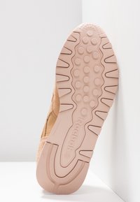 Sula av en ljusrosa mockaskor från Reebok med texturerat grepp och logotyp, placerad vertikalt mot vita hörnväggar.