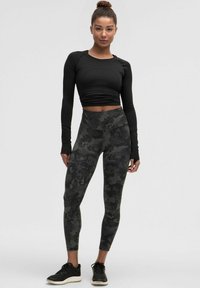 Crni dugih rukava cropped top uparen s tajicama visokog struka s tamnim kamuflažnim uzorkom. Nositi s crnim sportskim tenisicama.