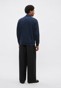 Tmavomodrá mikina s golierom, dlhými rukávmi a voľným strihom, doplnená širokými čiernymi nohavicami a jednoduchými čiernymi topánkami.