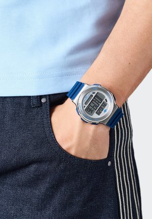 Reloj digital con una correa de silicona azul y caja plateada, con una gran pantalla digital y acentos azules. Llevado en una muñeca cerca de un bolsillo de mezclilla.