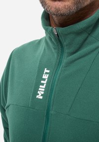 Grüne Fleecejacke mit hohem Kragen, ausgestattet mit einem halben Reißverschluss und strukturiertem Stoff. Die Marke "MILLET" ist auffällig in Weiß aufgedruckt.