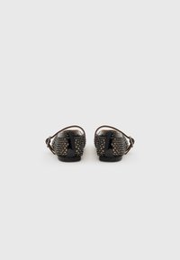 Sandales vernies noires avec un dos tissé texturé orné de petites rivets dorés, accompagnées d'une bride ajustable à la cheville. Design à talon bas.