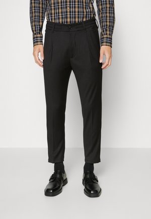 Mężczyzna noszący czarne spodnie na miarę, czarne skórzane eleganckie buty oraz brązowo-czarną koszulę w kratę z długim rękawem, stojący na białym tle.