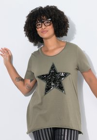 Camiseta verde oliva con una gran estrella de lentejuelas negras. Mangas cortas, escote redondo y un corte holgado. El detalle texturizado de las lentejuelas resalta.