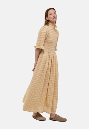 Femme portant une robe maxi vichy jaune avec des manches courtes à volants et un décolleté, associée à des chaussures marron à enfiler, debout sur fond blanc.