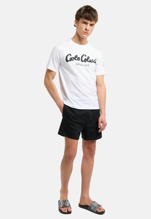 Jonge man draagt een wit Carlo Colucci T-shirt, zwarte korte broek en patroon slippers, staat met één hand in de zak.