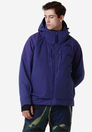 Giovane uomo con giacca blu con cappuccio e pantaloni a fantasia mimetica, guarda verso sinistra con le mani rilassate ai lati.