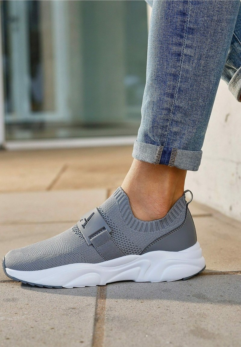 Scarpa da ginnastica grigia in maglia con cinturino elastico e suola bianca, dotata di punta rotonda e design in tessuto testurizzato, posizionata su una superficie piastrellata.