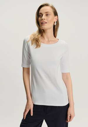 Kobieta z włosami blond do ramion, ubrana w biały top z krótkim rękawem i ciemne spodnie, stojąca na tle jednolitego jasnego tła.