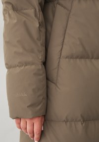 Gevoerde bruine parka met een gladde textuur, voorzien van een ruime zak en subtiele branding op de mouw. Close-up van de arm en de stofdetails.