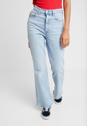 Person iført lyseblå jeans med høj talje, rød ærmeløs top og sort-hvide sneakers, stående mod en ensfarvet lys baggrund.