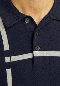 Námornícky polo tričko s pletenou textúrou, s dvoma čiernymi gombíkmi a bielymi horizontálnymi pruhmi ako akcentom. Detailný pohľad na golier a oblasť hrude.
