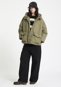 Chaqueta acolchada de color verde oliva con un diseño de acolchado, grandes bolsillos y capucha, superpuesta sobre un suéter a rayas, combinada con pantalones negros de pierna ancha y zuecos.