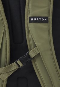 Bandoulière de sac à dos vert olive avec quincaillerie noire et texture tissée, arborant une étiquette avec "BURTON" en lettres blanches. Le fond est noir.