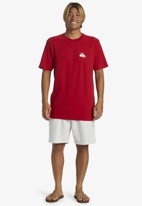 Camiseta de algodón roja con cuello redondo y mangas cortas, adornada con un logo gráfico blanco en el lado izquierdo del pecho. Combinada con pantalones cortos de color gris claro.