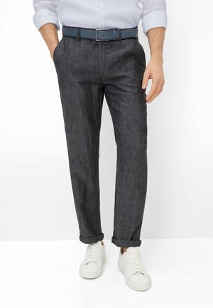 Man draagt donkergrijze opgerolde broek, witte sneakers, blauwe riem en een wit hemd met lange mouwen, met één hand in de zak.