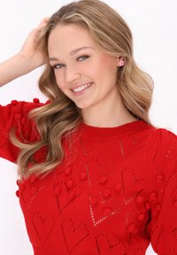 Rød strikket sweater med hjerte mønstre og pom-pom detaljer, med en rund halsudskæring. Modellen har bølget hår og bærer runde lyserøde blomsterøreringe.