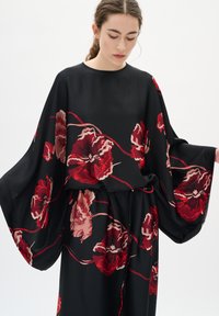 Rochie neagră cu flori, cu mâneci largi, având flori mari roșii și roz și contururi delicate, realizată dintr-un material neted și fluid.