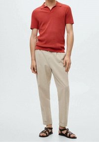 Polo rosso a maniche Corte con colletto, abbinato a pantaloni tapered beige chiaro e sandali neri con cinturini.