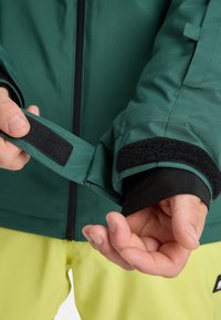 Grüne wasserdichte Jacke mit schwarzen Klettverschlüssen an den Bündchen, sichtbaren Nähten und einem Reißverschluss vorne. Hand passt das Bündchen mit schwarzem Innenfutter an.