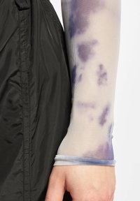 Črno najlonasto zapestje jakne v kombinaciji s svetlo, prosojno mrežasto rokavno manšeto s vijoličastim tye-dye vzorcem in prilegajočim zapestnim trakom.