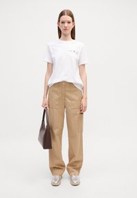 T-shirt bianco in cotone con logo, pantaloni cargo beige a vita alta, e sneakers argentati. La modella tiene una borsa marrone scuro. Design minimalista.