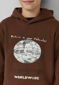 Brązowa bluza z kapturem z grafiką globu i sceny na lotnisku, z białym napisem "Uwierz w swój potencjał" oraz "WORLDWIDE."