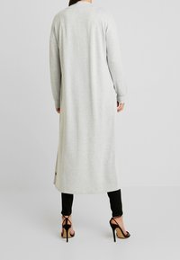 Lange grijze cardigan van zachte stof, met een rechte snit en geribbelde manchetten, gecombineerd met zwarte broek en sandalen met hoge hakken.