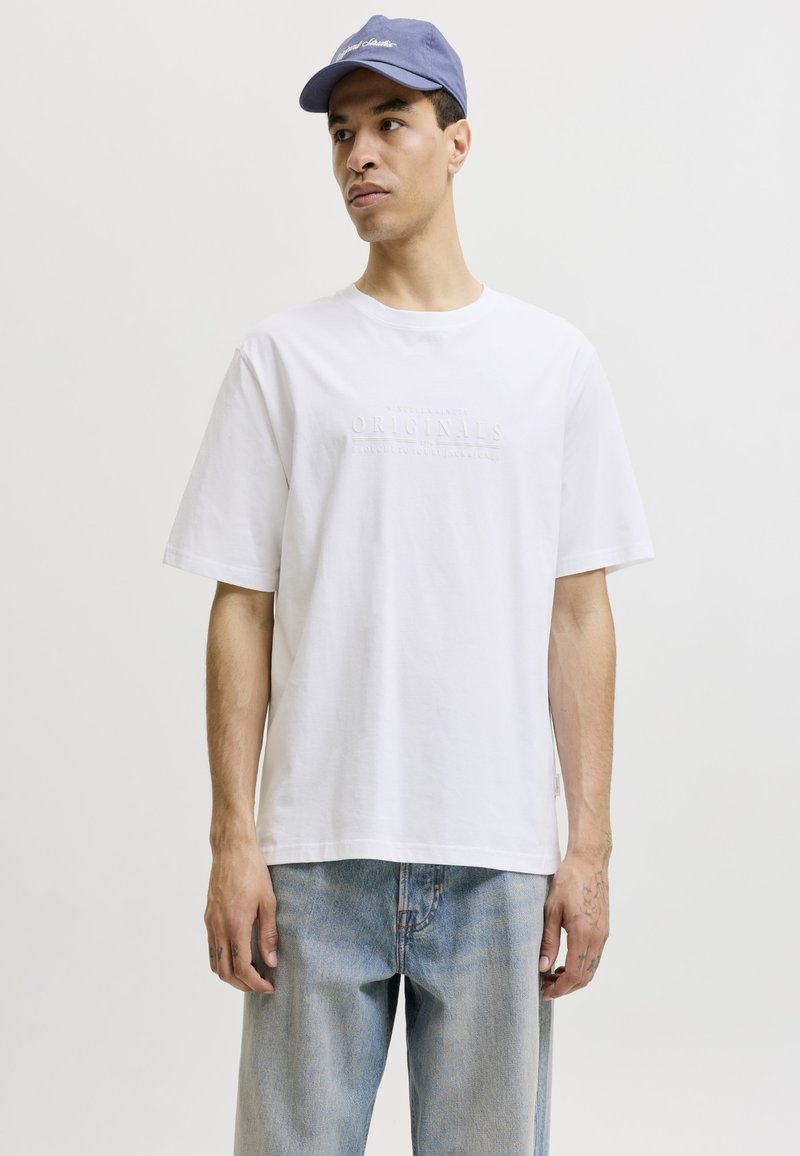 Άνδρας που φοράει απλό λευκό φαρδύ T-shirt, ανοιχτό μπλε τζιν και μπλε καπέλο baseball, στέκεται μπροστά σε απλό φωτεινό φόντο.