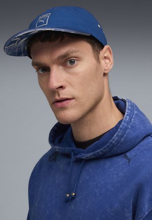 Blauer Cap mit einem gemusterten Schirm, Logo auf der Vorderseite; getragen mit einem passenden blauen Hoodie mit einer strukturierten Oberfläche und Kordeln.