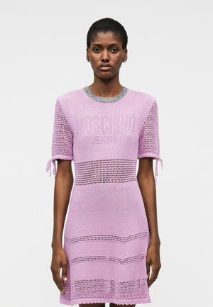 Femeie purtând o rochie tricotată liliac cu modele perforate, mâneci scurte legate la manșete și inscripția texturată „Moschino Jeans” pe piept.