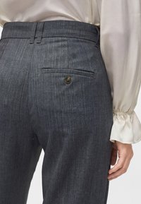 Gros plan d'une personne portant un pantalon gris taille haute avec un bouton sur la poche arrière et un chemisier crème à manches longues avec des poignets à volants.