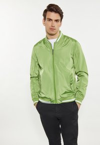 Giacca bomber verde con finitura lucida, polsini e orlo a coste, chiusura con zip e tasche laterali, abbinata a pantaloni neri.