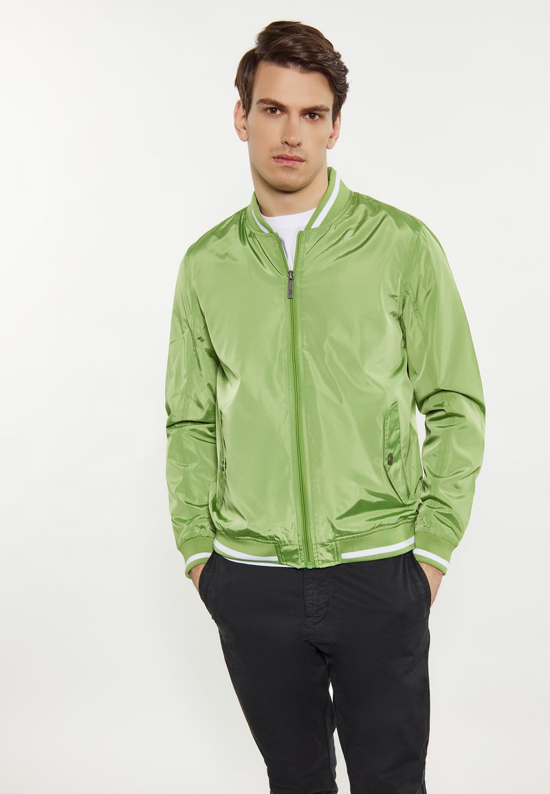 Giacca bomber verde con finitura lucida, polsini e orlo a coste, chiusura con zip e tasche laterali, abbinata a pantaloni neri.