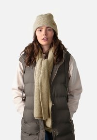 Groene puffer vest over een beige trui, gecombineerd met een gestructureerde beige sjaal en een lichtbeige gebreide muts. Gladde en geïsoleerde materialen door het geheel.